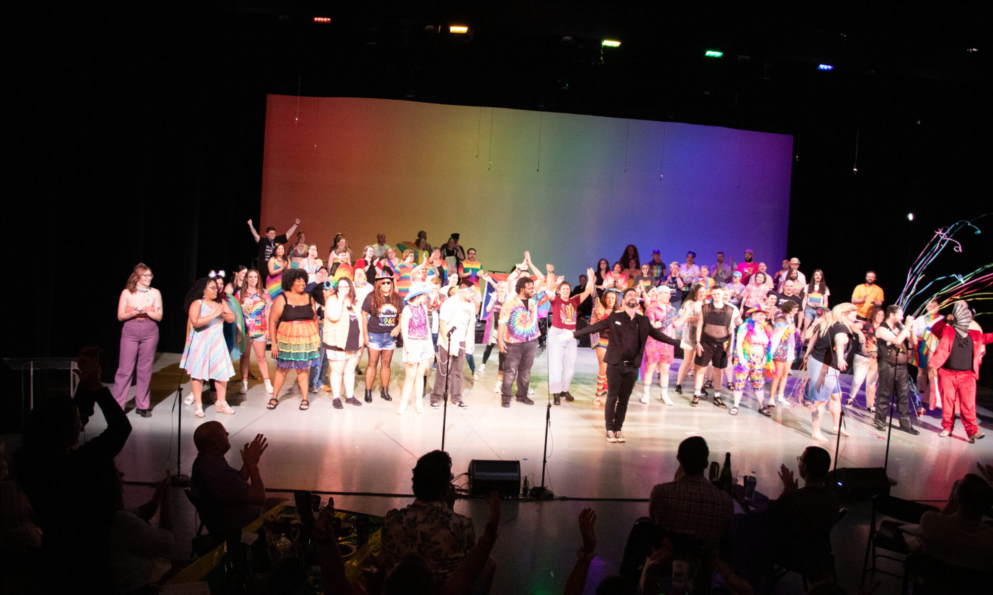 A large group of singers take a bow after a performance in front of a rainbow backdrop.